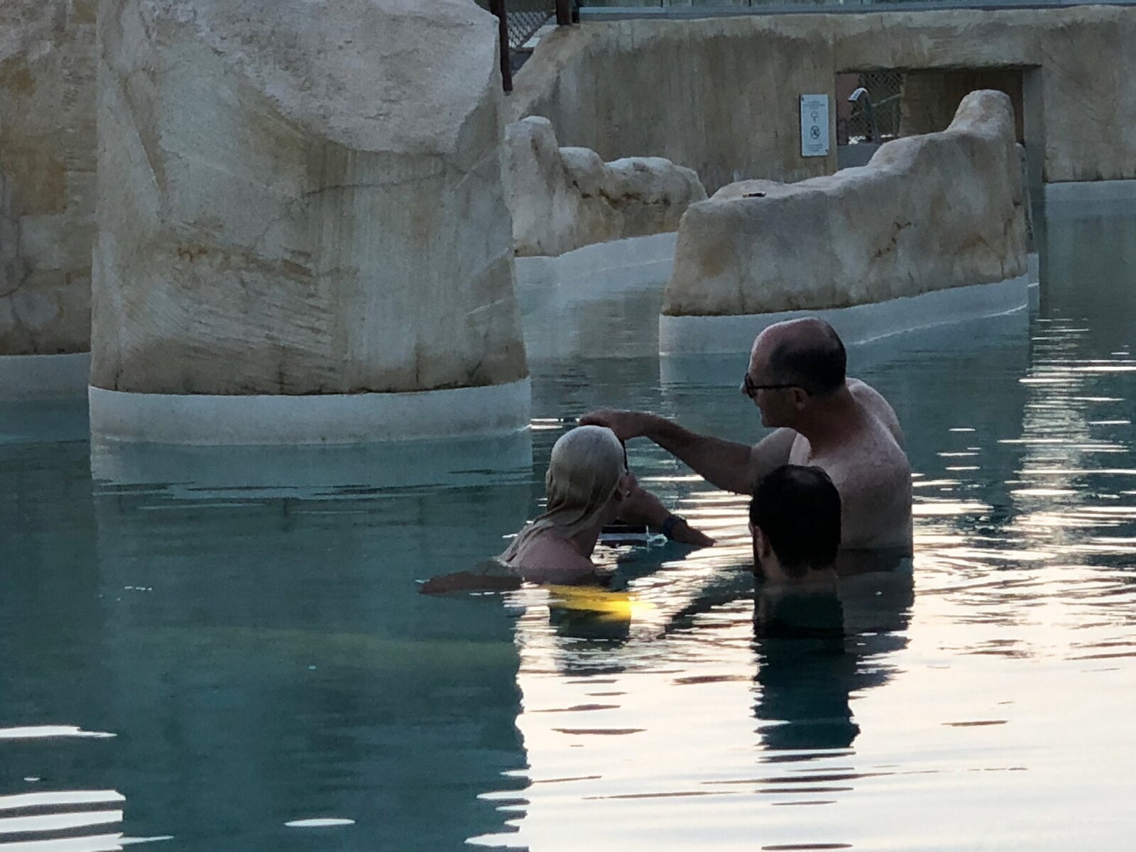 Three crew members stand in an indoor pool between artificial rock formations, gathered around partially submerged equipment.