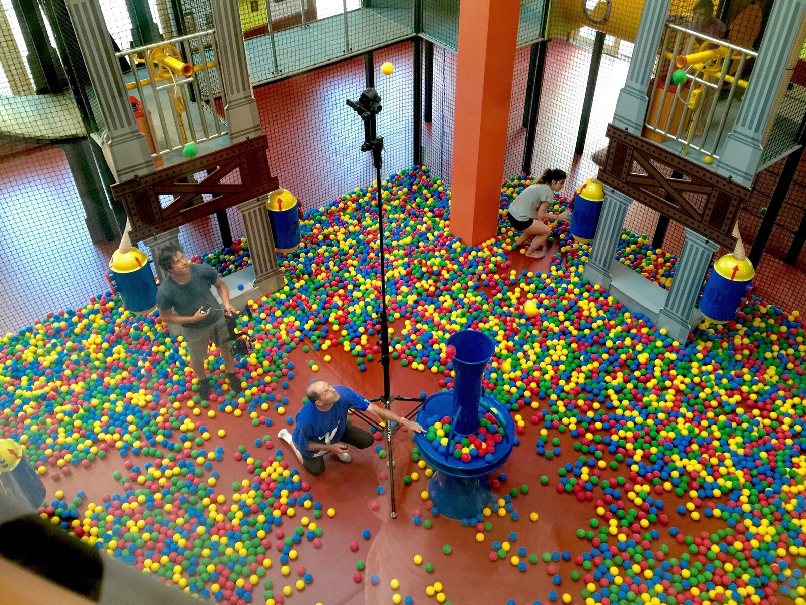 Three crew members work around a tall camera rig in a brightly coloured indoor ball pit.