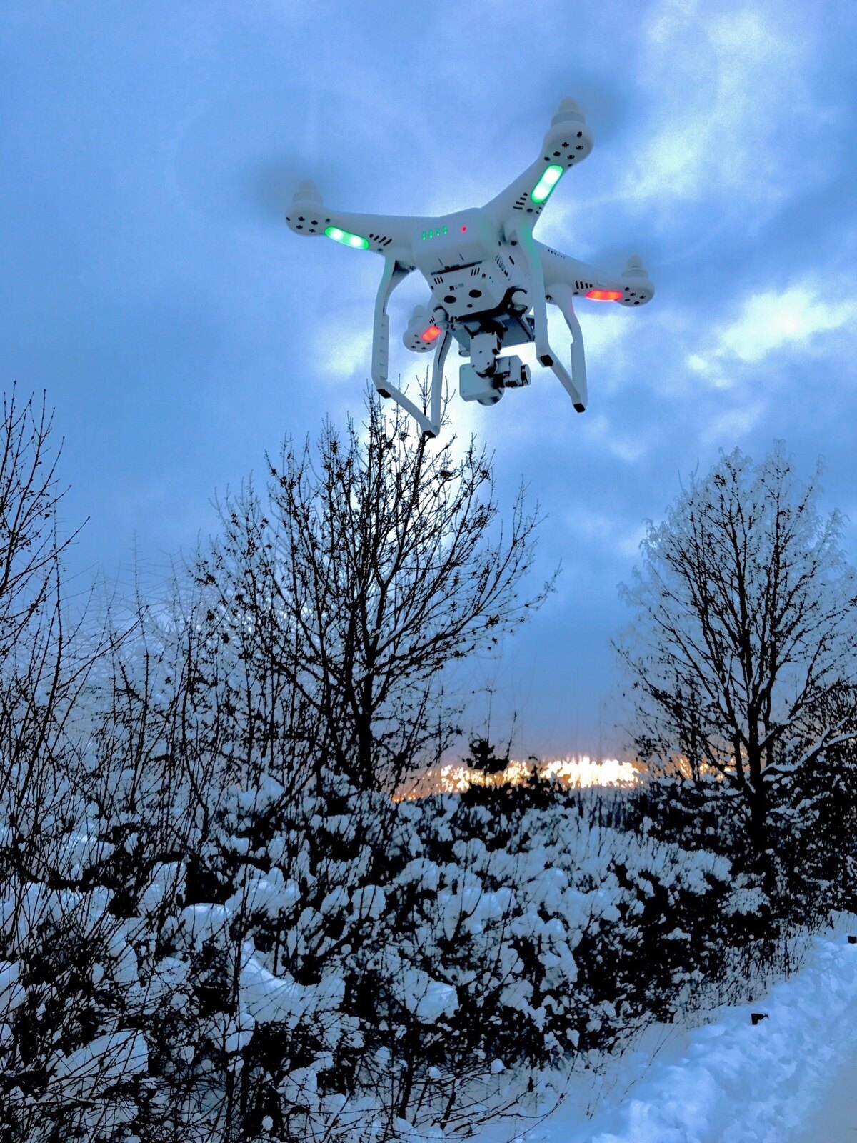 White camera drone hovering above snow-covered trees under a cloudy sky.