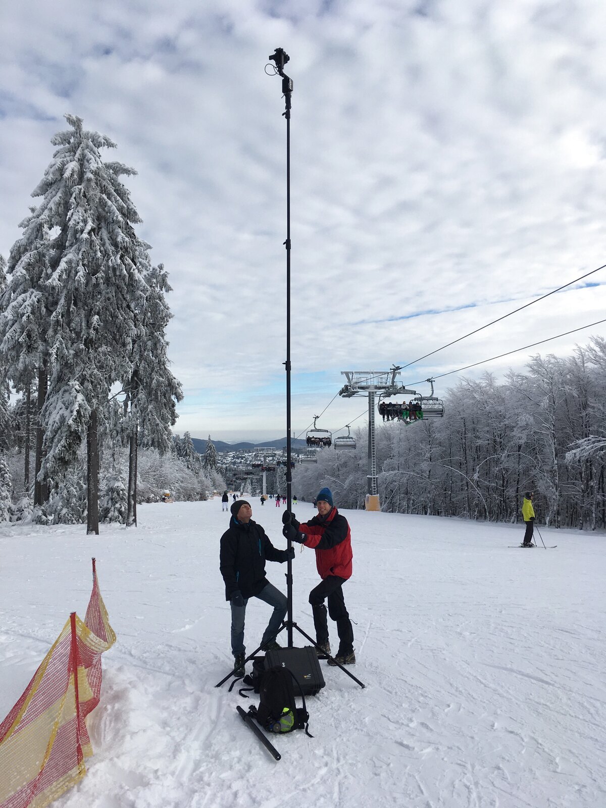 Two crew members hold a tall camera mast on a snowy ski slope beneath chairlifts.