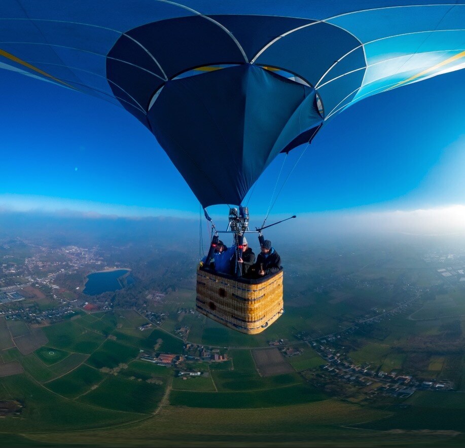 Blue hot-air balloon carrying passengers above fields and woodland.