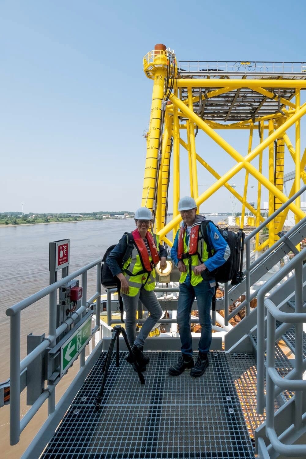 Two crew members in hard hats, high-visibility vests and life jackets stand on a steel platform beside a yellow industrial structure and river.