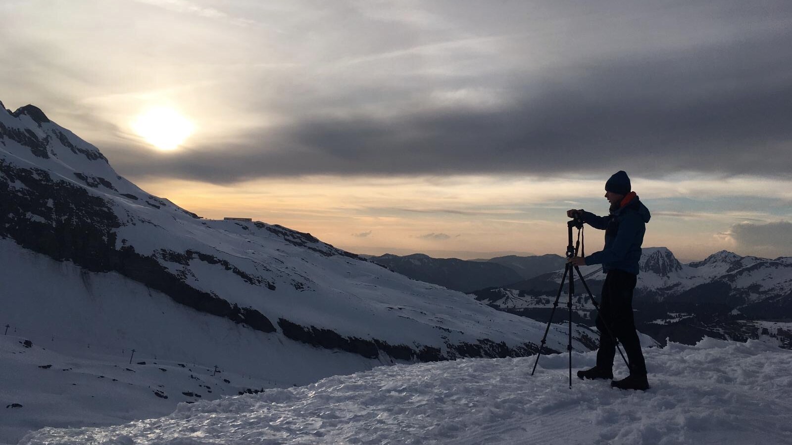 Camera operator adjusting a tripod on a snowy mountain at sunset.