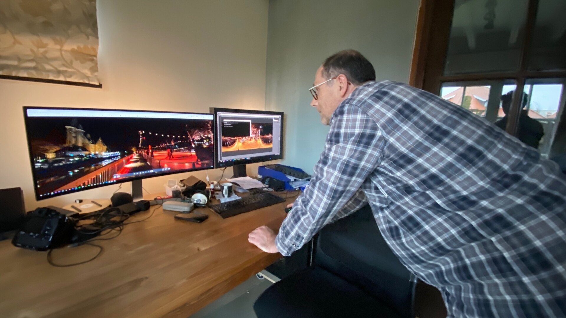 Person leaning toward two monitors displaying a brightly lit nighttime waterfront scene, with camera gear on the desk.