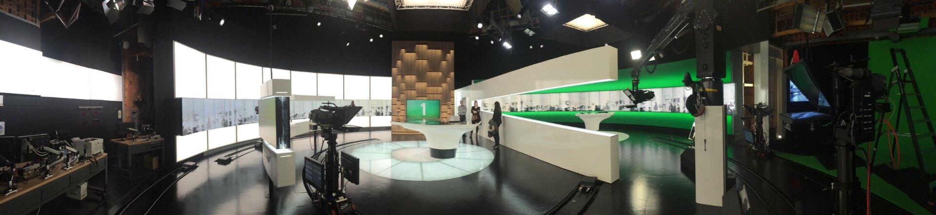 Wide panoramic view of a television studio with broadcast cameras, overhead lights, a green screen and crew.